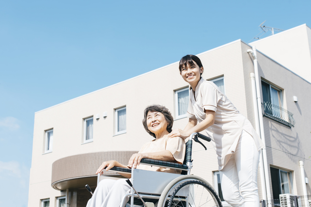 施設の前で微笑む介護職の女性と車椅子の高齢者